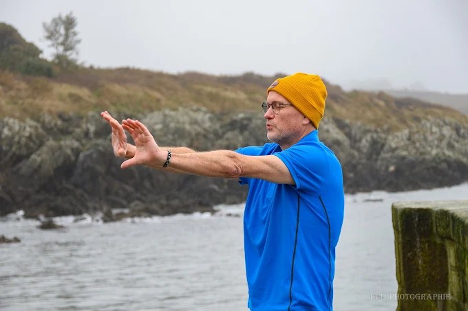 Alain Percevault guide un exercice de respiration au bord de la mer, les bras tendus devant lui